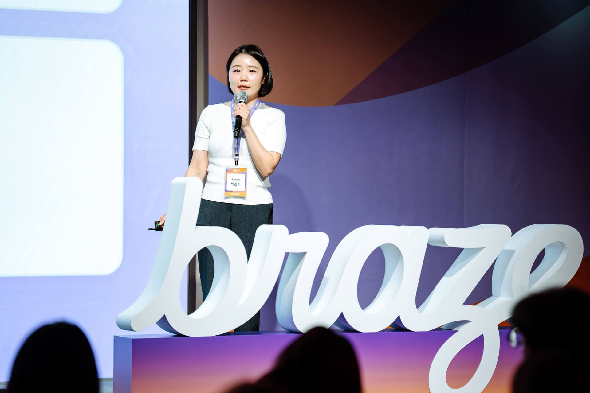 A woman with a microphone speaks on a stage with a large white "braze" logo.