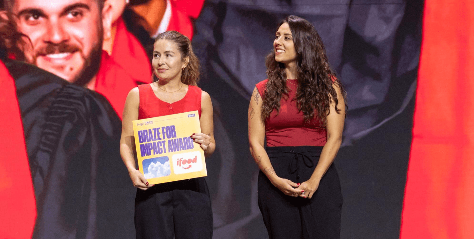 Two women on a stage, one holding a yellow "BRAZE FOR IMPACT AWARD" plaque with the iFood logo.