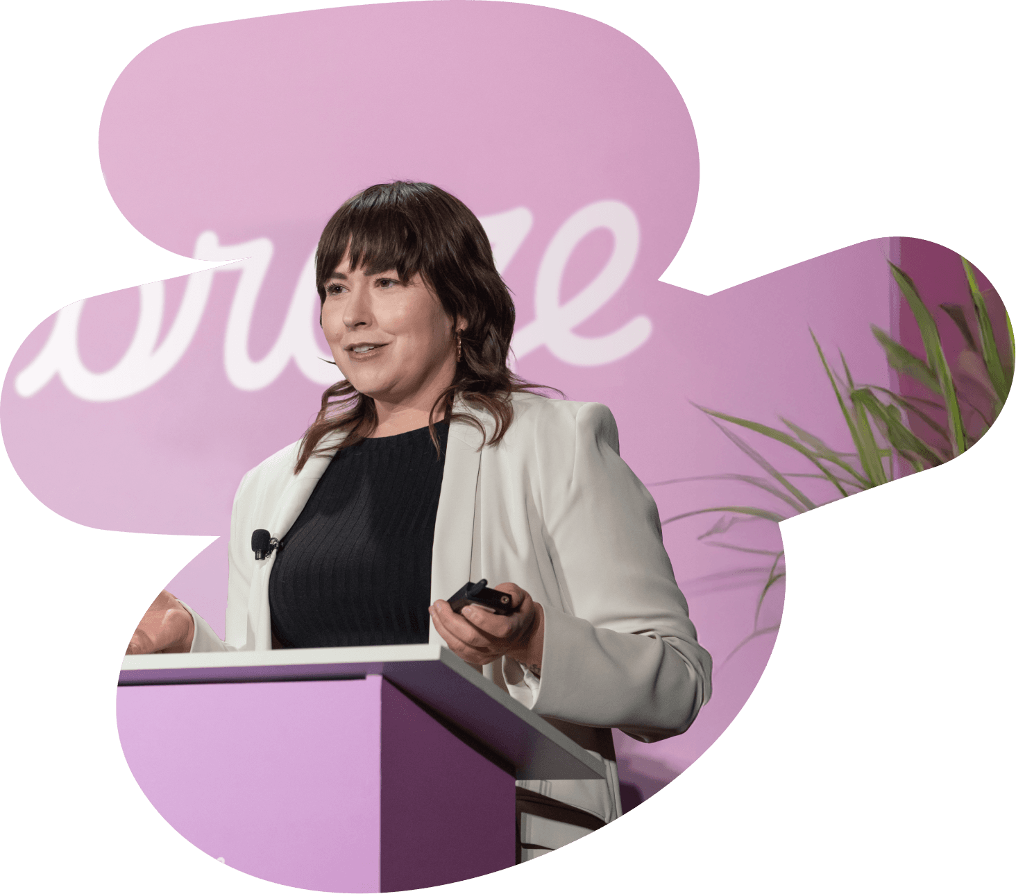 A woman in a white blazer speaks at a purple podium, holding a clicker, against a pink background with the word "embrace."
